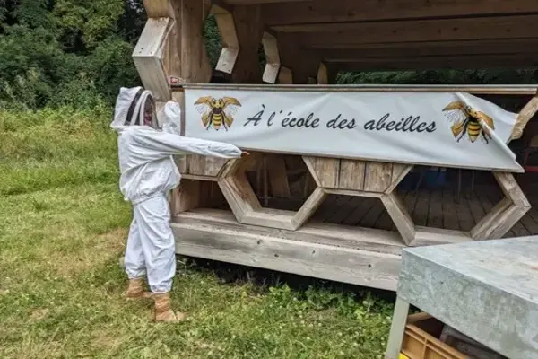 Une personne avec un costume de protection blanc devant une banderole sur un batiment en bois avec des formes d'alvéoles de ruche, où il est écrit "À l'école des abeilles".