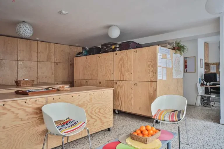 Photo du salon de la bagagerie. Au premier plan, 2 fauteuils blancs sont autour d'une table basse où est posée une corbeille de fruits contenant des oranges. Au fond à gauche, on voit les casiers des accueillis, et à droite, un bout du bureau d'accueil