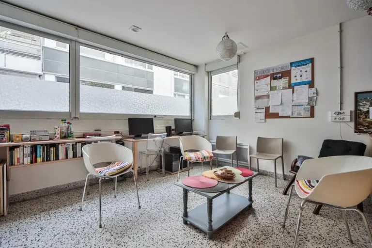 Photo du salon de la bagagerie. On y voit des fauteuils autour d'une table basse où est posée une corbeille qui contient des viennoiseries. Au fond à gauche, on voit, sous les fenêtres une petite bibliothèque, et 2 ordinateurs en libre-service.